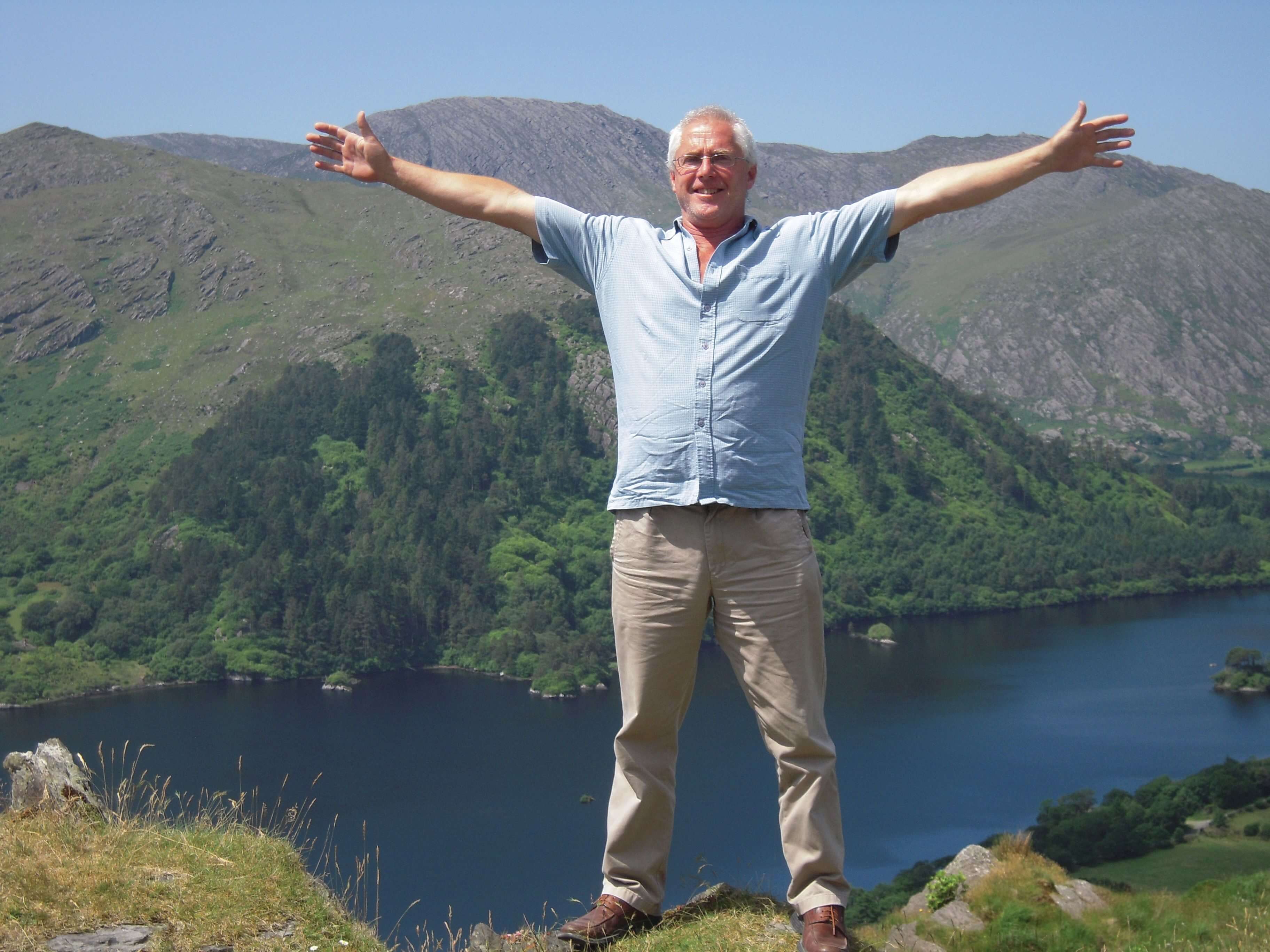 Male Tour Leader wearing blue shirt and beige cargo pants. His arms are spread wide as he stands in front of large green hills and body of water.