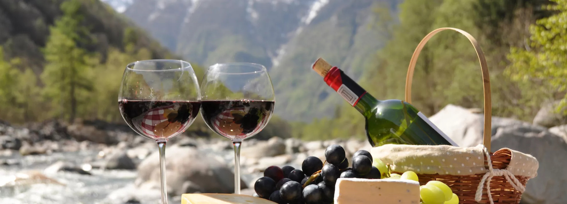 a picnic basket and two glasses of red wine, cheese and grapes in the foreground and a Swiss mountain scene in the background