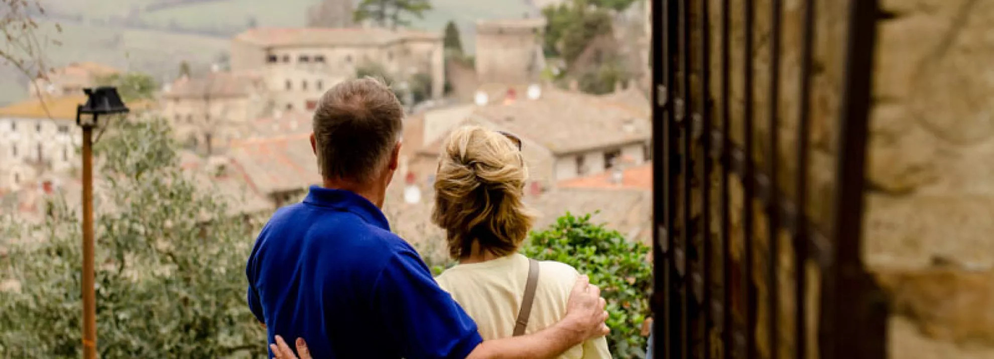 middle aged couple facing with backs to camera and arms around one another looking out over an Italian village skyline