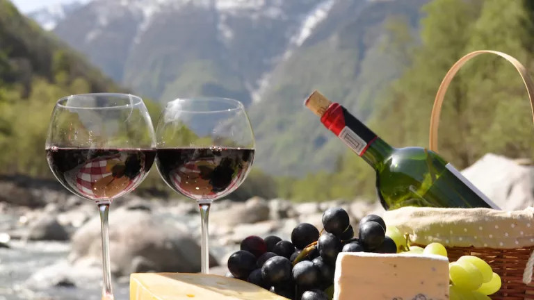 a picnic basket and two glasses of red wine, cheese and grapes in the foreground and a Swiss mountain scene in the background