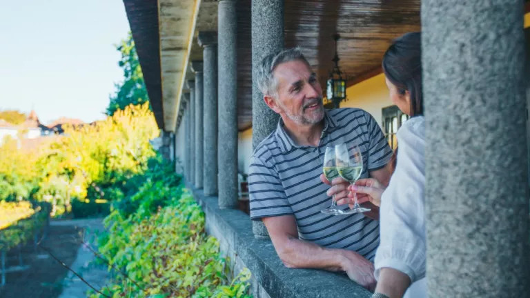 middle aged man and woman clinking glasses of white wine in a portico overlooking a leafy garden