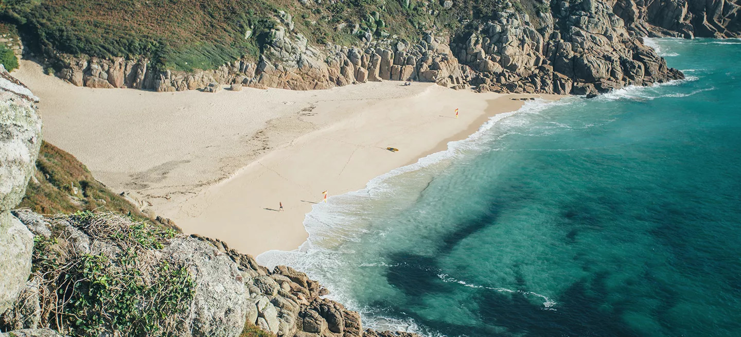 Aerial view of Porthcurno Beach, Cornwall, showing turquoise waves, sandy shore, and surrounding cliffs.