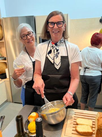 Two mature women wearing glasses and black aprons are smiling at the camera. One whisks an egg whilst the other holds a glass of white wine.