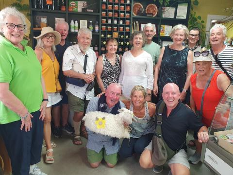 A small group of mature people in a sheep farm shop, the middle man kneeling as he holds a fake sheep in his arms